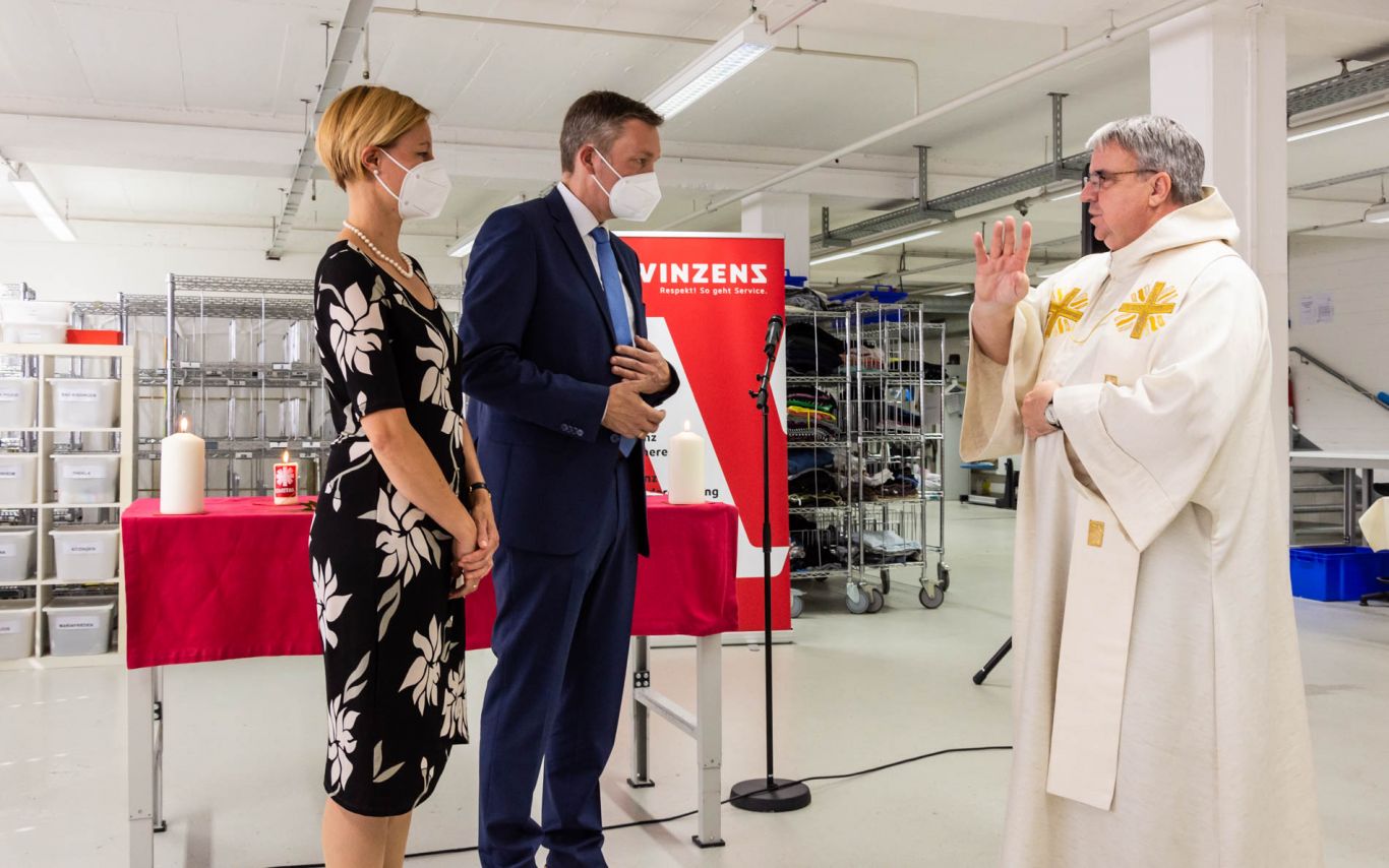 Domkapitular segnet Christoph Vogel in einer Werkhalle von Vinzenz vor einem Tisch mit Kerzen und roter Tischdecke.