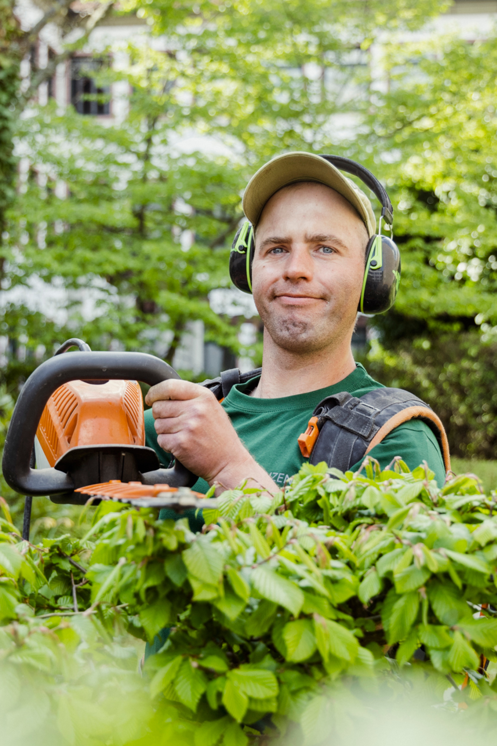 Junger Mann mit Schallschutz-Kopfhörern und elektrischer Heckenschere im Grünen.