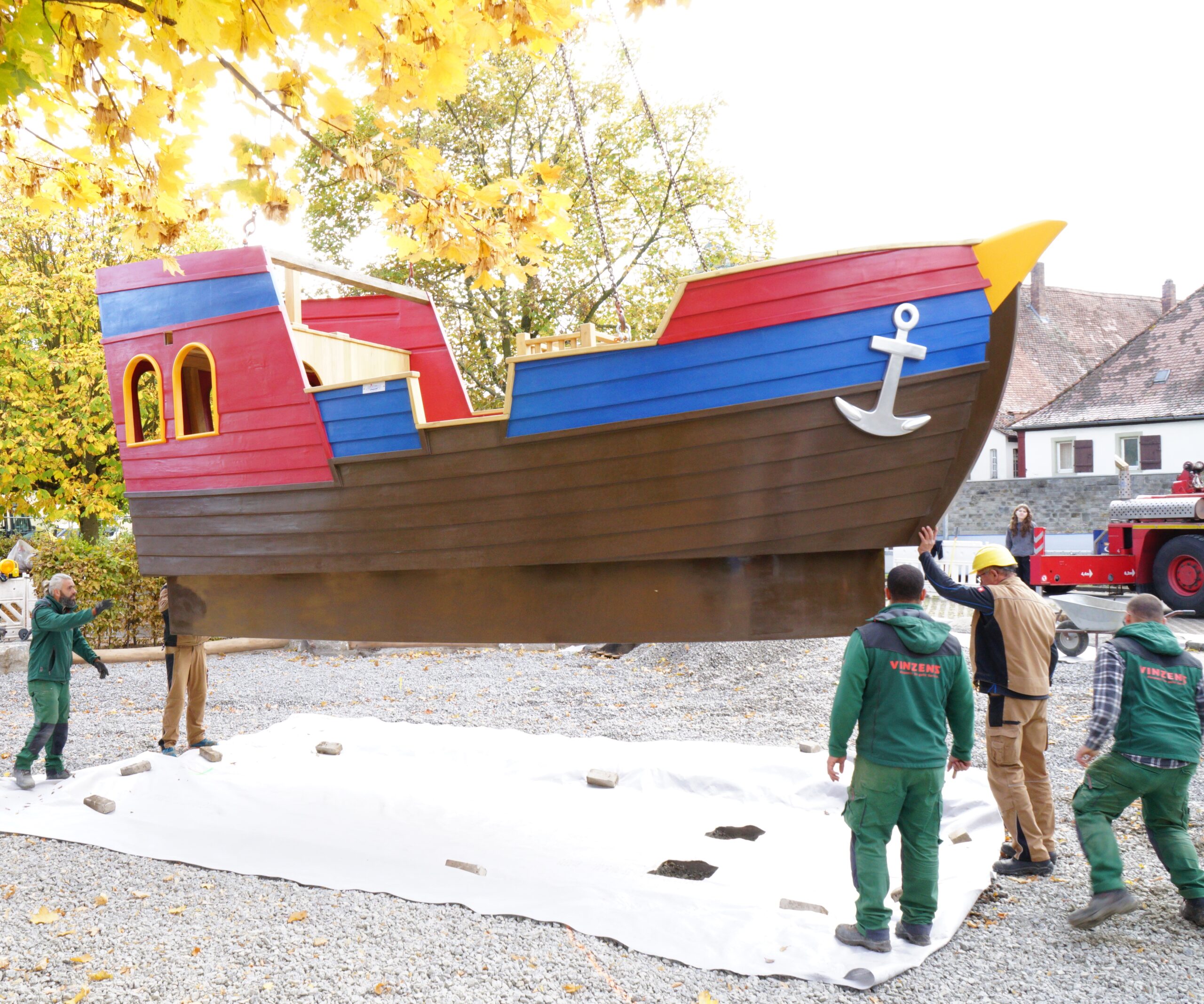 Das Bild zeigt mehrere Kollegen von Vinzenz GaLa Bau beim Platzierung des Aktivschiffs der Playmobil Stiftung. Das XXL-Schiff schwebt am Haken des Schwerlastkrans.