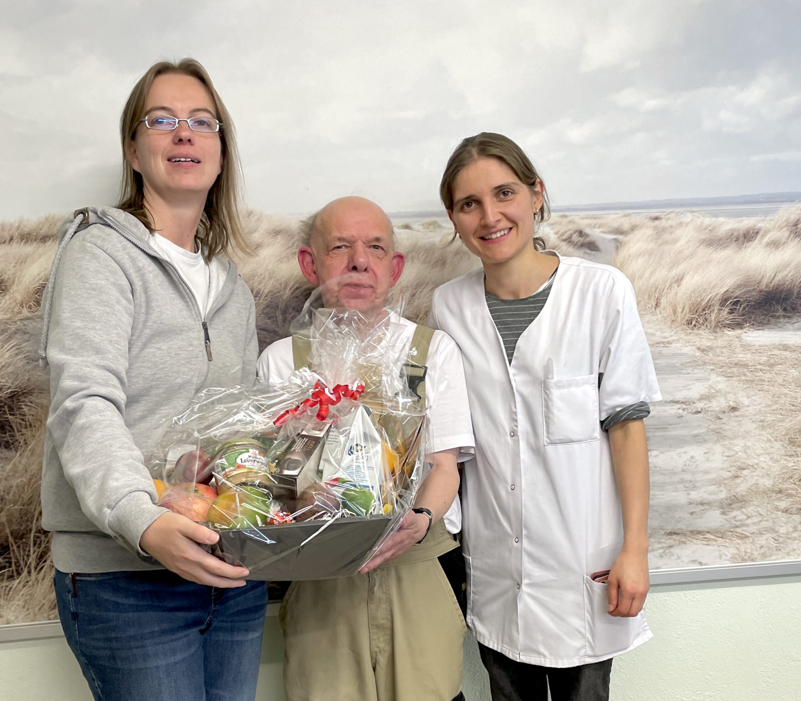 Im Bild von links nach rechts: Kathrin Tatgenhorst-Arnold, stellvertretende Betriebsleiterin Vinzenz Wäscherei; Leo Dietz; Daniela Basha, Betriebsleiterin Vinzenz Wäscherei. Sie stehen vor einem großformatigen Bild, auf dem eine Stranddüne zu sehen ist. Frau Tatgenhorst überreicht Herrn Dietz einen Präsentkorb.