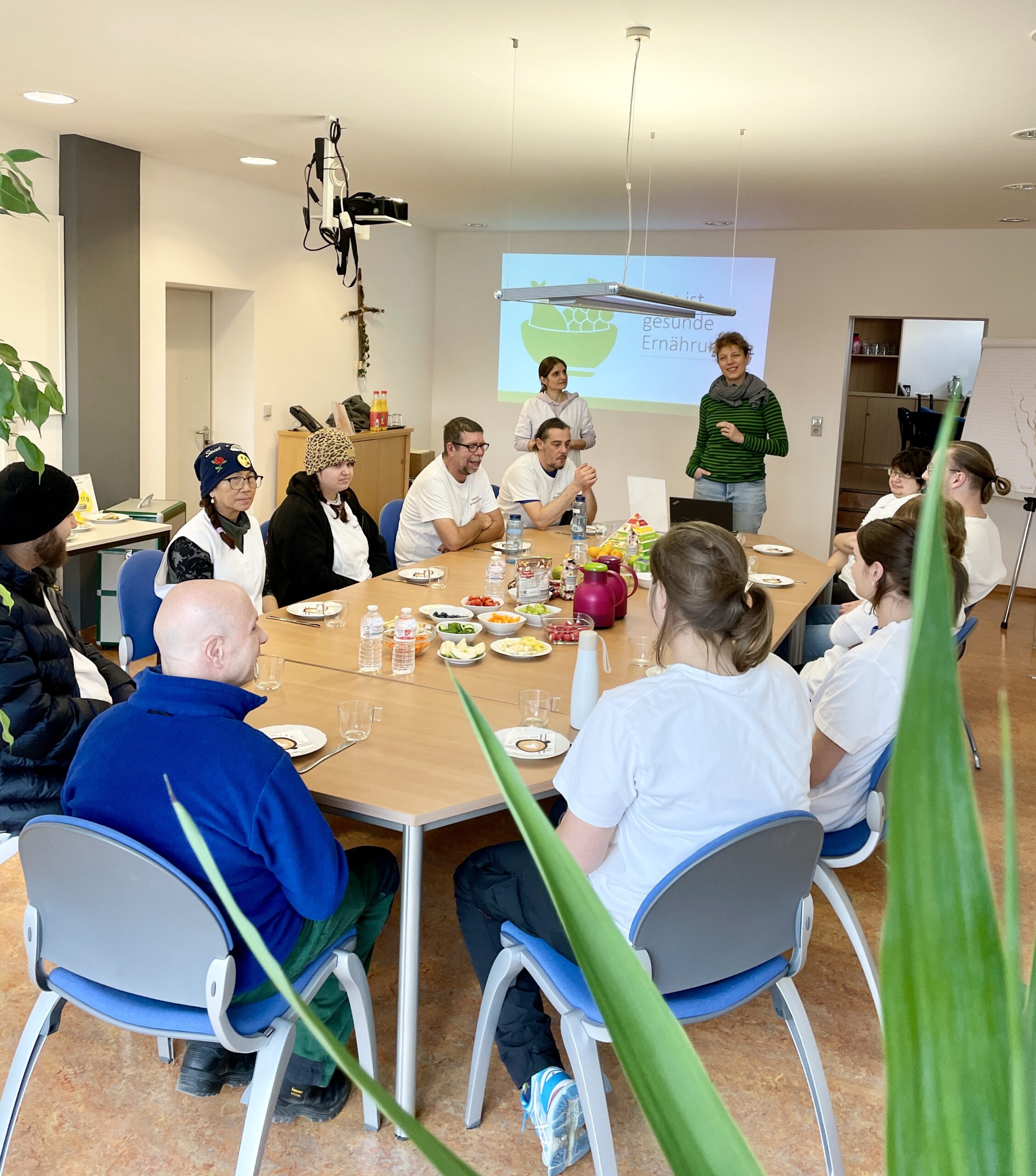 Um einen großen Frühstückstisch im Besprechungsraum sitzen interessierte Kolleg*innen aus der Wäscherei. Frau Reeg und Frau Basha stehen am Kopfende des Tisches und präsentieren. Im Hintergrund ist eine Chart mit der Überschrift "Was ist gesunde Ernährung" zu sehen.