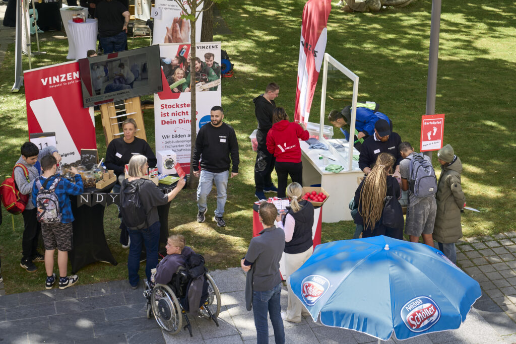 Auf dem Bild ist der Messestand von Vinzenz Würzburg zu sehen. Mitarbeitende unterhalten sich angeregt mit Interessent*innen
