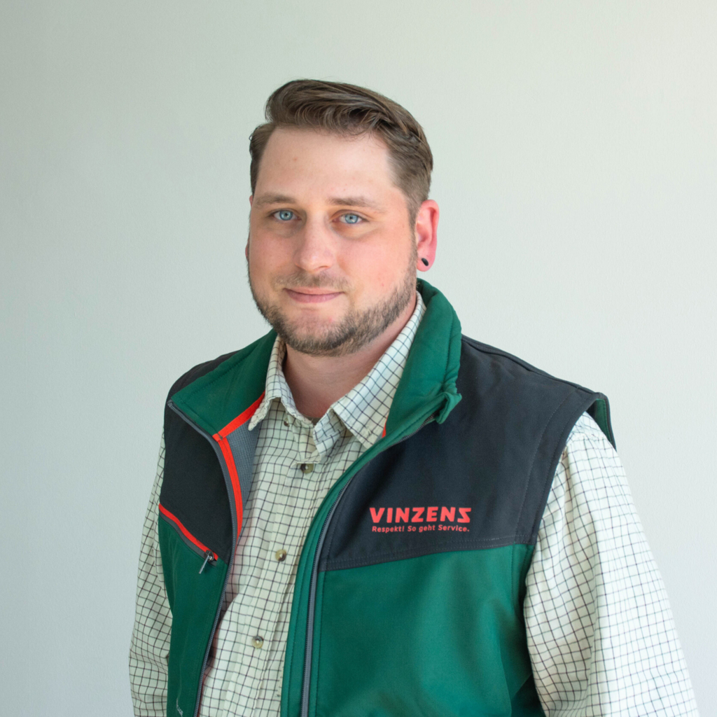 Portraitbild von Pascal Schäfer. Er lächelt in die Kamera. Blonde, kurze Haare, gestutzter Bart, kariertes Hemd. Darüber trägt er eine grüne GalaBau-Weste mit Vinzenz-Logo