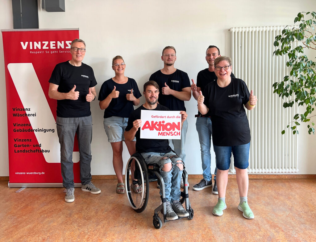 Ein Gruppenbild von Kolleg*innen - alle lächeln mit Daumen hoch. Der Kollege im Vordergrund im Rollstuhl hält ein Schild "gefördert durch die Aktion Mensch" hoch.