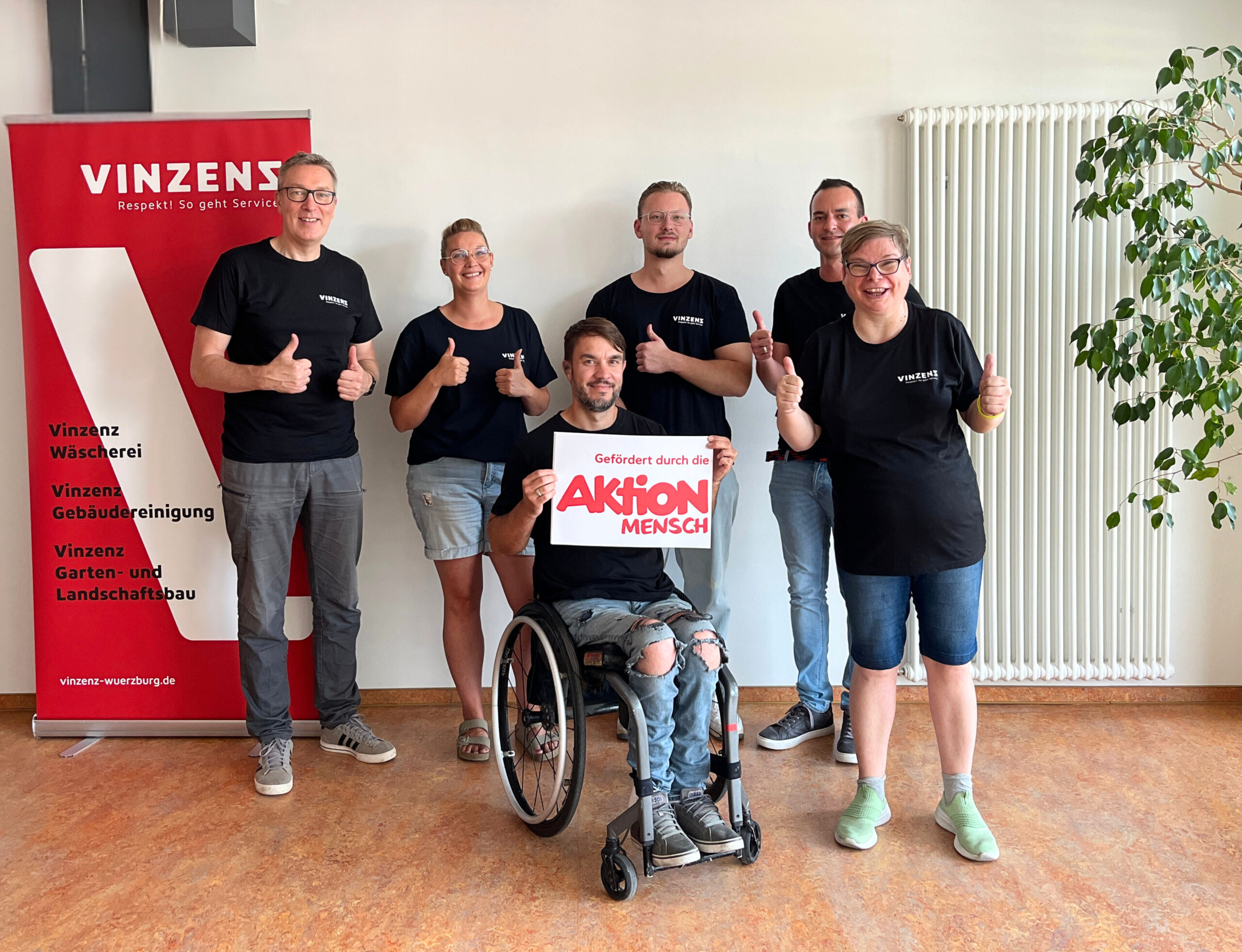 Ein Gruppenbild von Kolleg*innen - alle lächeln mit Daumen hoch. Der Kollege im Vordergrund im Rollstuhl hält ein Schild "gefördert durch die Aktion Mensch" hoch.