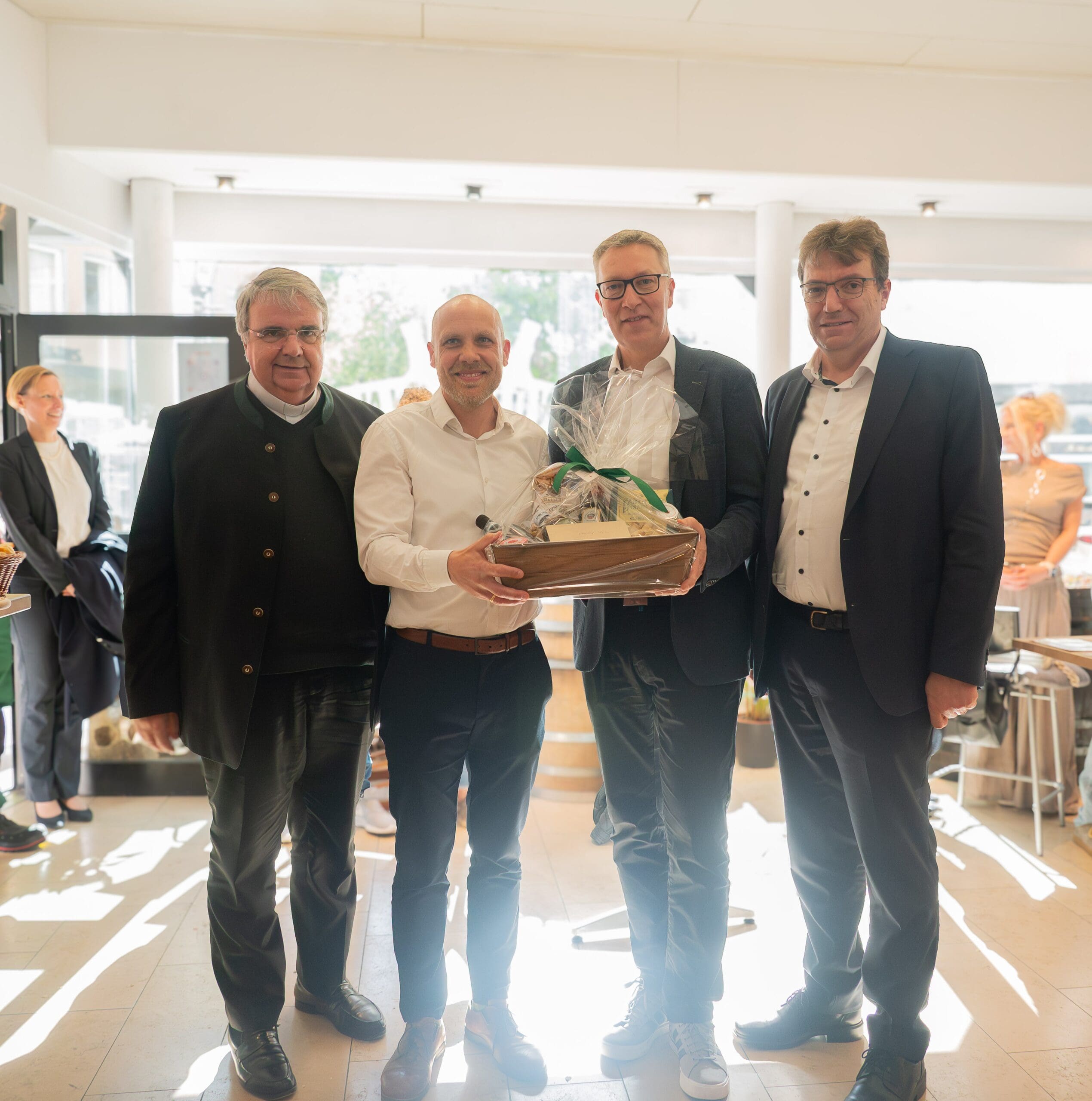 Domkapitular Bieber, Sperrle, Vogel und Düchs stehen lächelnd in einem hellen Innenraum nebeneinander. Sperrle und Vogel halten gemeinsam einen großen, in Folie verpackten Geschenkkorb. Alle tragen Business-Kleidung oder festliche Garderobe.