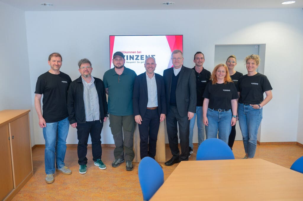 Gruppenbild des Teams von Vinzenz Würzburg mit Generalvikar Dr. Jürgen Vorndran in einem Besprechungsraum. Geschäftsführer Georg Sperrle und der Generalvikar stehen zentral, umgeben von Mitarbeitenden. Im Hintergrund ist das Logo von Vinzenz Würzburg zu sehen.