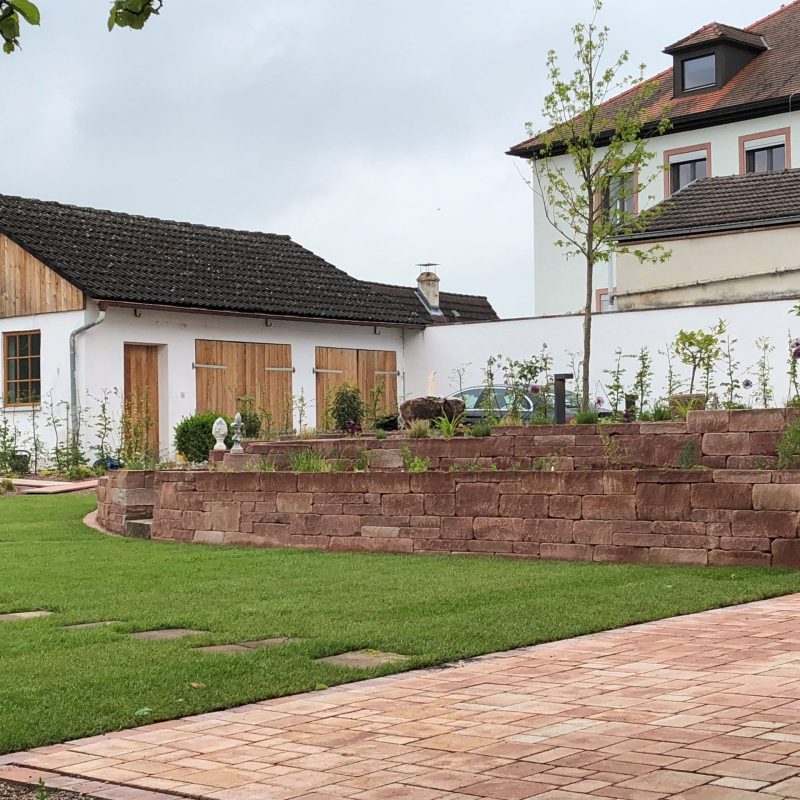 Rötliche Natursteinmauern und gepflasterter Platz in Gartenanlage.