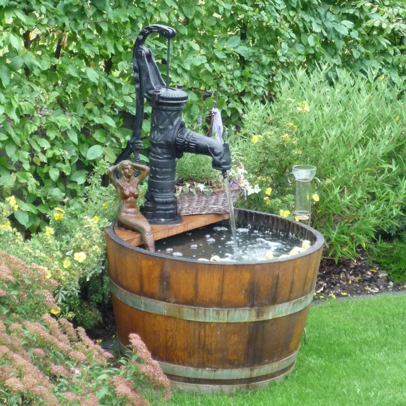 Schwengel-Pumpe mit nackter Frauenfigur auf Holzfass im Garten.