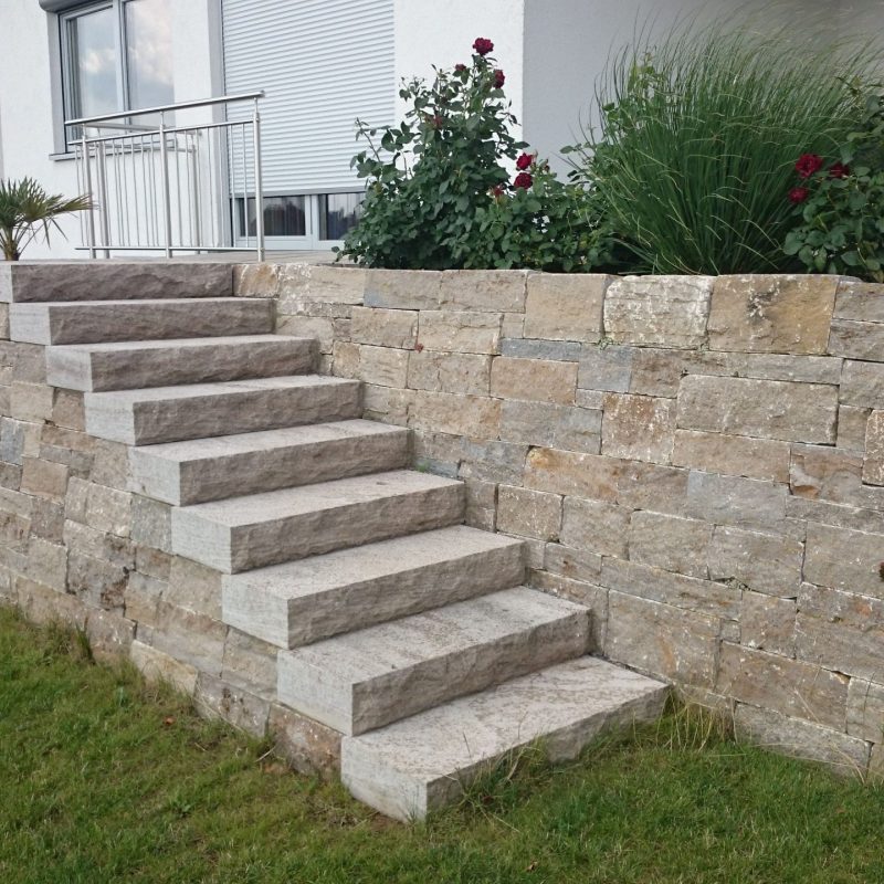 Treppe vor Natursteinmauer führt auf die Terrasse von Einfamilienhaus.