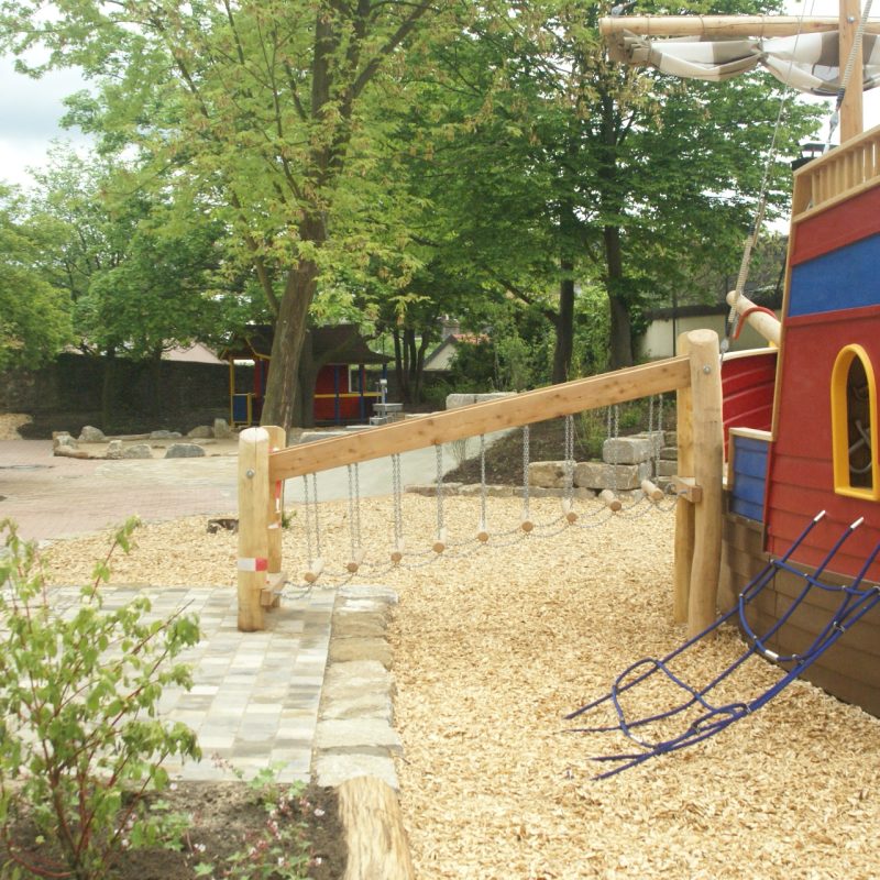 Seilbrücke mit einzelnen Balken führt zu Holzschiff auf Spielplatz.